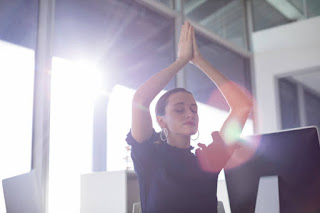 Woman seated at her desk practicing yoga, raising her hands in a prayer position, illustrating mindfulness and relaxation in adaptive yoga.