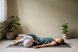 Person lying in Savasana pose on a yoga mat, using a bolster under knees, relaxing in a dimly lit room, peaceful and restful atmosphere