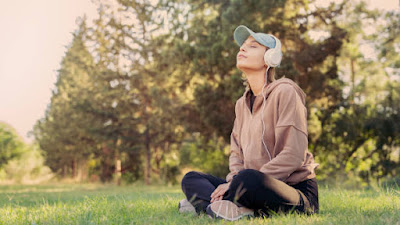 A woman sitting cross-legged in a grassy field, listening to music with headphones, embodying mindfulness and a sense of oneness with nature, inspired by formless Jhana experiences.