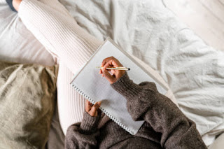 A person writing in a journal while seated cross-legged on a comfy bed.