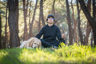 A teenage boy sitting cross-legged outdoors, eyes closed, practicing deep breathing with a peaceful expression in a natural setting.