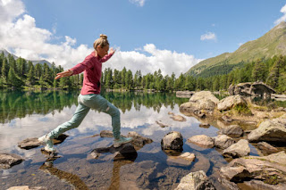Person stepping across rocks in a serene lake surrounded by mountains and trees, representing courage and progress in life’s journey.