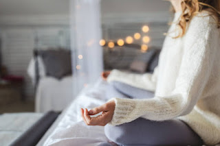 A woman in a meditative pose on her bed, wearing a cozy sweater, surrounded by warm, calming light—perfect for balancing emotions and setting intentions for the day.