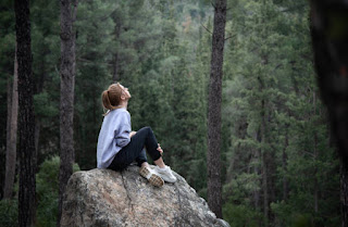 A woman sitting on a large rock in a dense forest, looking upward peacefully, surrounded by tall pine trees.