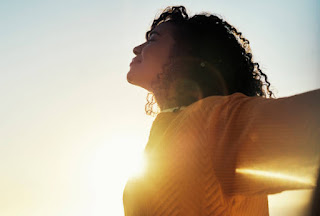 A woman with curly hair in a yellow top, basking in the sunlight with her arms outstretched, radiating joy and positivity.