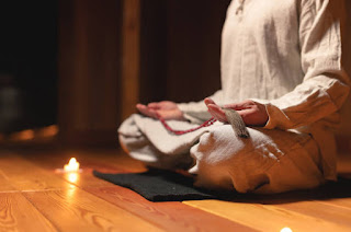A person sitting cross-legged on a wooden floor, holding prayer beads, surrounded by candles in a dimly lit, peaceful room.