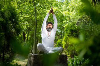A man in white sitting on a rock, raising his arms overhead in a meditative posture, surrounded by lush greenery, symbolizing connection with nature and spiritual grounding.