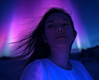 A person with a dreamy expression stands within a glowing aurora of purple, blue, and pink hues.