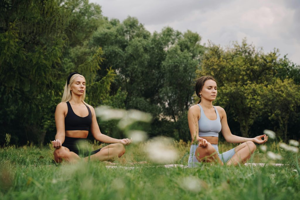Two friends work together to manage their pain symptoms with yoga in a beautiful clearing surrounded by nature. Tuning into their breath.